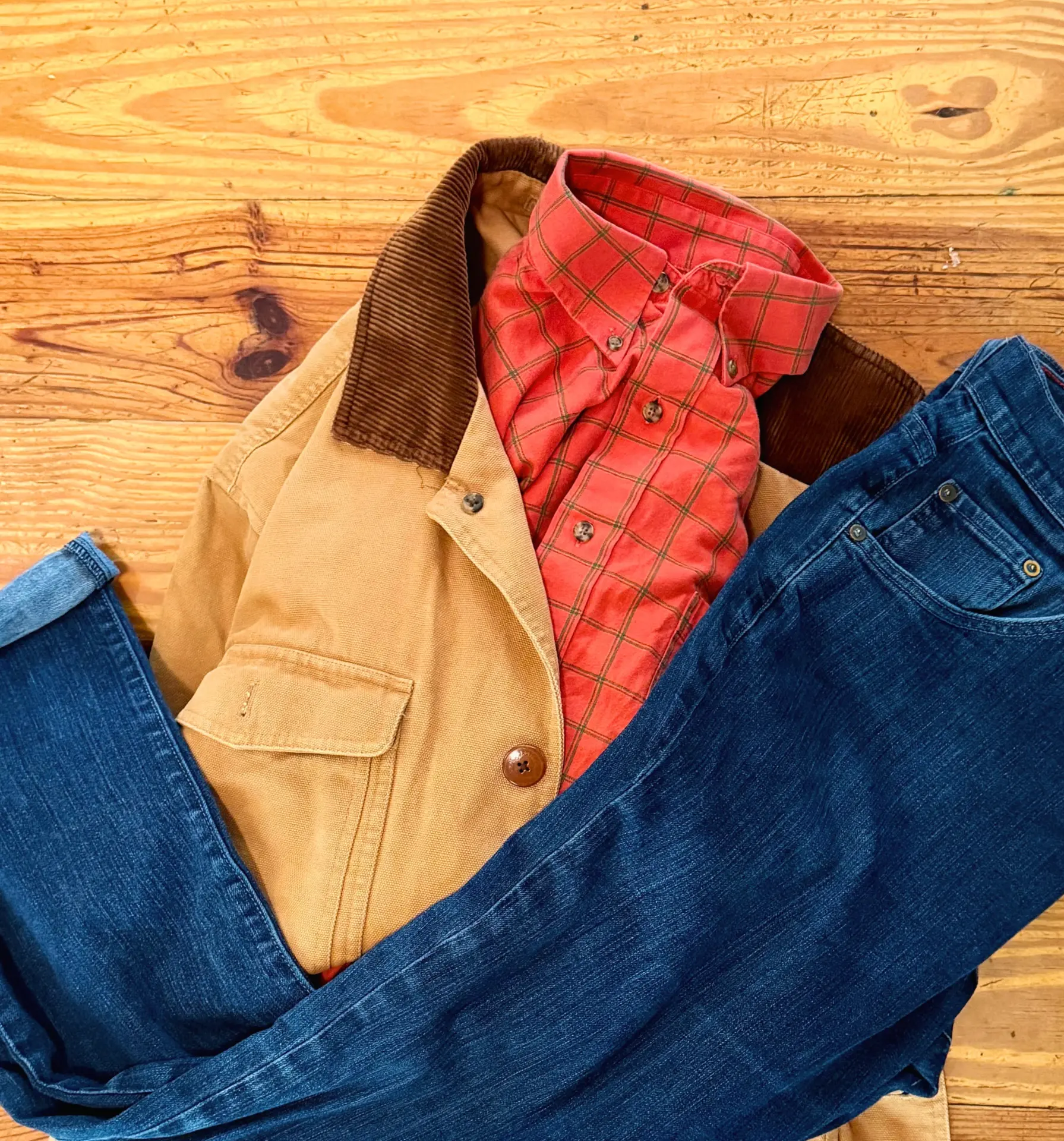 A tan canvas jacket with a brown corduroy collar, red plaid shirt, and blue jeans arranged on a wooden floor.