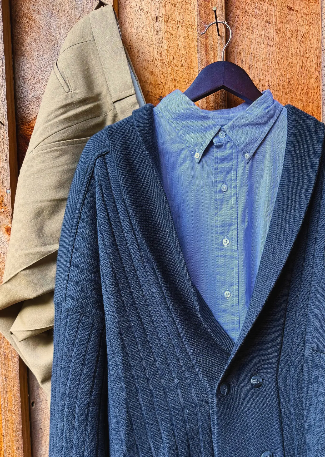 A navy shawl cardigan layered over a blue button-down shirt, hanging with khaki trousers against a wooden wall.