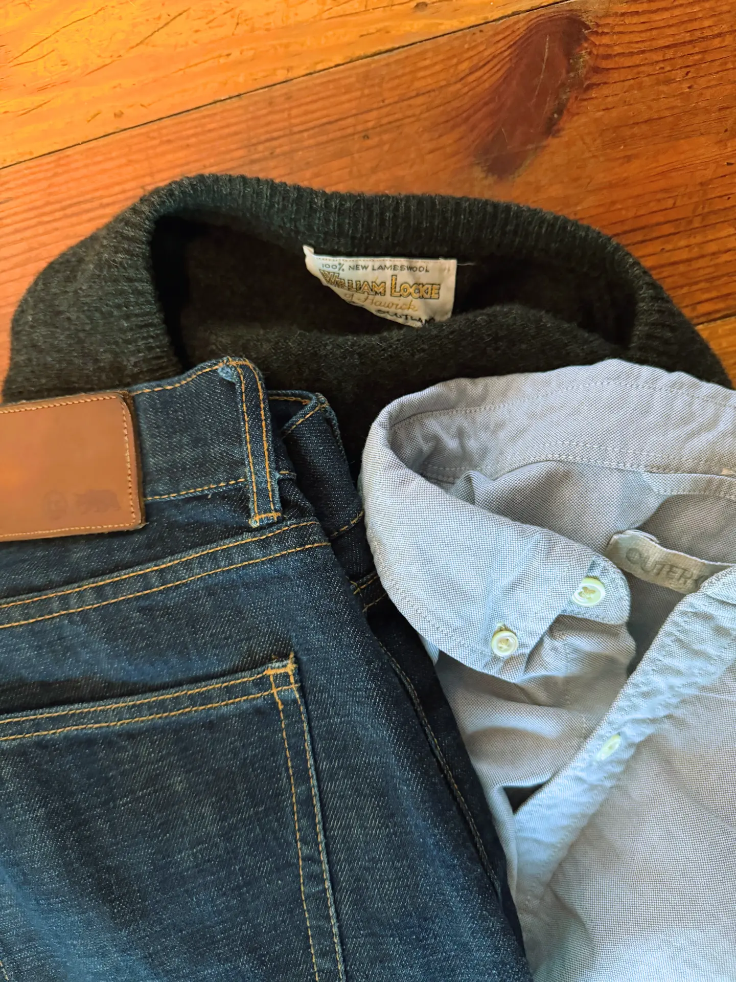 Close-up of a William Lockie lambswool sweater label beside a pale grey button-down shirt and dark raw-denim jeans.